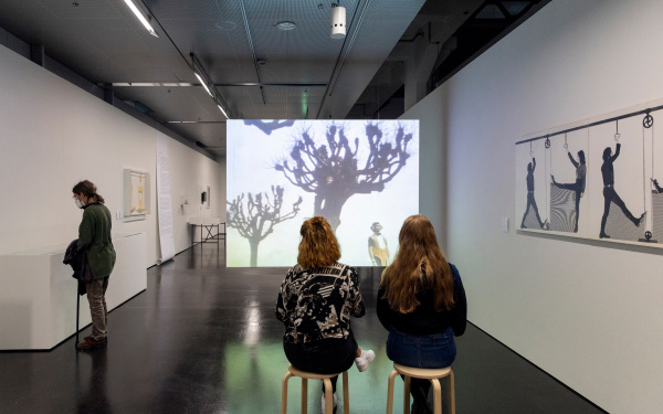 Three young women in the exhibition space. Two sit on stools in front of a film screen on which bare trees can be seen. An abstract drawing hangs on the wall to the right. On the left wall are boxes into which the third woman looks.