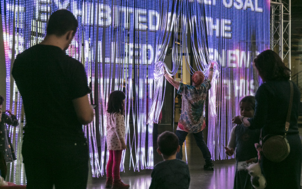 A woman steps through a curtain of light sticks onto which many bright colours and lettering are projected.