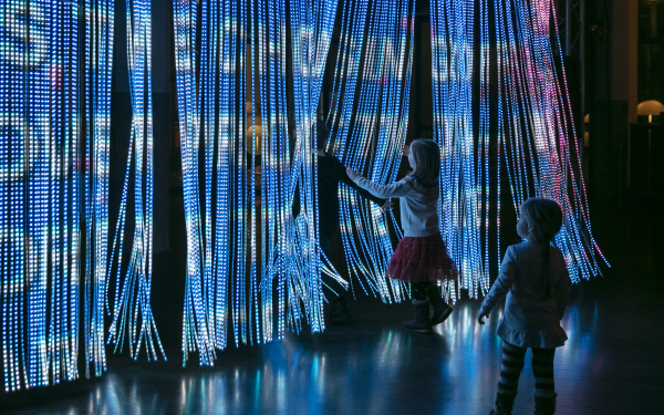 Kinder treten durch einen Vorhang aus Leuchtstäben auf die viele bunte Farben und Schriftzüge projektiert werden.