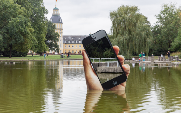 You can see the shore of a lake. A very large hand with a smartphone is sticking out of the lake. The display of the smartphone is a mirror in which the viewers can see themselves on the shore.