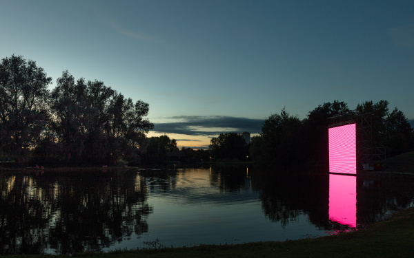 A large square area can be seen, which seems to stand on the surface of a lake in the dark of the night. On the surface the flickering of a screen can be seen. 