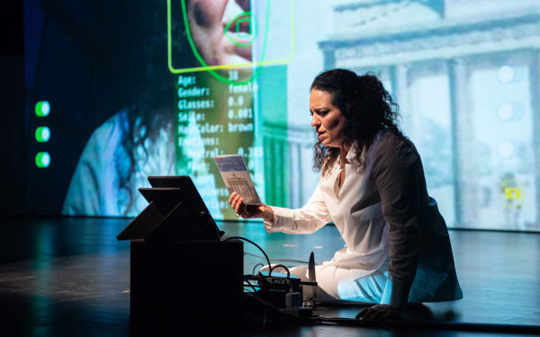 Noa Frenkel is sitting on stage in front of a laptop. Behind her there is a large projection of her, which comes from the laptop's webcam.