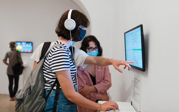 Eine Frau zeigt auf einen Bildschirm. Mit der anderen Hand hält sie eine Computermaus. Sie trägt Kopfhörer.