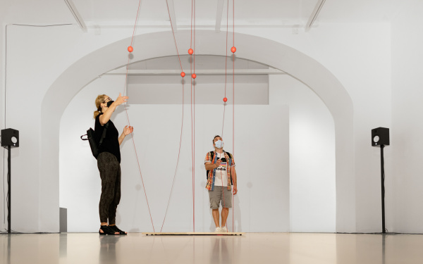 Ein Mann und eine Frau stehen vor einem Kunstwerk, das aus Seilen besteht, welche zwischen der Decke und dem Boden gespannt sind. Die Besucher ziehen die Seile auseinander.