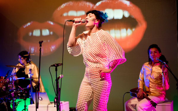 Die Sängerin der Band Chicks on Speed singt leidenschaftlich ins Mikrofon, hinter ihr Schlagzeugerin und Backroundsängerin. Auf einer großen Leinwand im Hintergrund sind drei große Münder zu sehen.