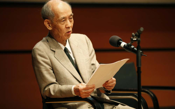 Hiroshi Kawano bei seinem Vortrag. Er trägt einen hellen Anzug, in den Händen hält er sein Manuskript.