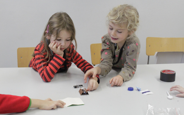 Two girls sitting at a table are building a small robot using a motor, batteries, the head of a toothbrush and other utensils.