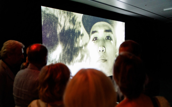 Das Bild zeigt eine Menschenmenge vor einem riesigen Bildschirm auf dem gerade ein asiatischer junger Mann mit Cappy zu sehen ist.