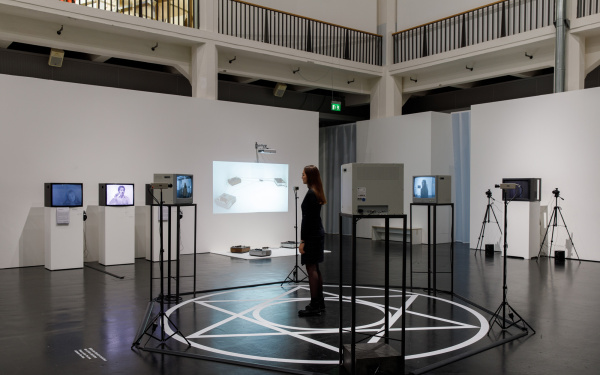 A woman is standing in a white hexagram on the floor. A circle is drawn in the center of the hexagram and around it. At the tips of the star there are three cameras and three screens, showing the woman, directed towards the centre.