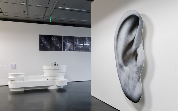 On the left, a white installation that looks like a futuristic bar. In the background black and white images of the universe. On the right half of the picture a wall with an oversized pressure of an ear.