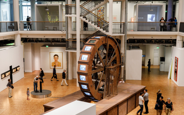 Ein riesiges Mühlrad aus Metall steht in einem Ausstellungsraum. Am Mühlrad befinden sich Röhrenbildschirme worauf Videos von Wasser abgespielt werden.