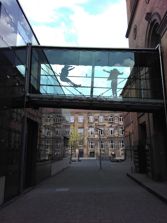 Das Bild zeigt den Blick in eine Gasse mit Sandsteinhäusern. Zwischen den Häusern sieht man eine schräg verlaufende Brücke, die mit Glas abgedeckt wurde. Auf der Brücke springen zwei Personen, die man durch das Glas sillhouettenhaft sieht..