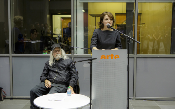 Links im Bild sitzt Ira Schneider, Medienkünstler, auf einem Stuhl und hört Daria Mille, Kuratorin am ZKM, bei ihrem Vortrag zu.