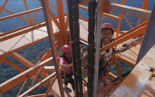 Zu sehen sind zwei Frauen mit Helmen auf ihren Köpfen, die grinsend an einem Stahlgerüst in die Höhe steigen.