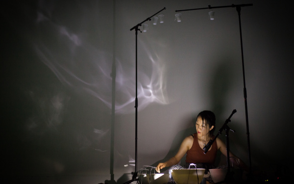 Tomoko Sauvage sitzt auf dem Boden, vor ihr stehen mit Wasser gefüllte Schalen. An der Wand sind Lichtreflexionen des Wassers zu sehen.