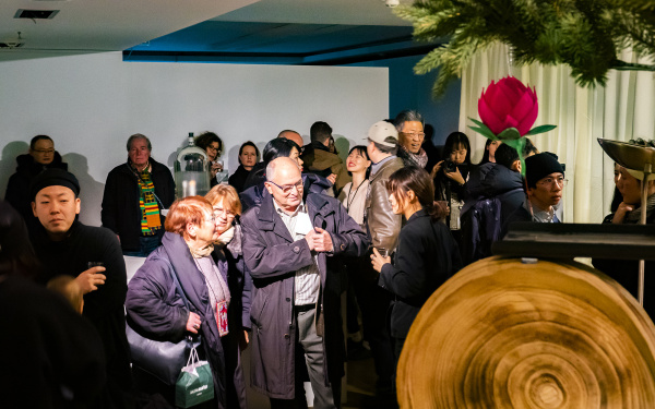 Zu sehen sind viele Menschen in der Galerie damdam in Berlin. Auf der rechten Seite des Bildes erscheint verschwommen ein Tannenzweig darunter ist eine große Rote Papierblüte zu sehen.