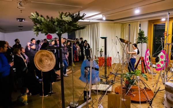 Zu sehen ist der Kulturraum Korea in Berlin während einer Performance. Auf der linken Seite stehen die BesucherInnen während auf der rechten Seite eine Performerin sich zwischen verschiedenen Soundobjekten bewegt.