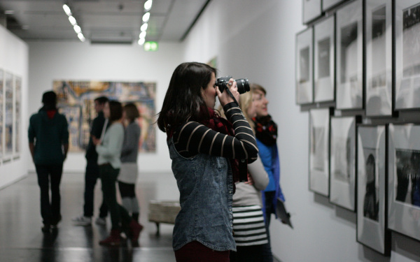 A woman is standing in the museum in front of numerous pictures on the wall. She is holding her camera and is making a picture from the pictures.