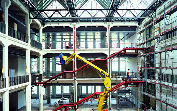 Das Foto zeigt eine historische Ansicht des Umbaus des alten MNK Hallenbaus. Ein imposanter großer gelber Bagger steht mitten in der Halle während im Hintergrund die Rohkonstruktion der zukünftigen Treppen des Ausstellungsraumes in rot hell leuchten.