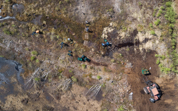 Eine Dronenaufnahme von oben, die auf eine Moorlandschaft blickt. Auf ihr arbeiten Menschen mit Müllsäcken, Baggern und Paletten.