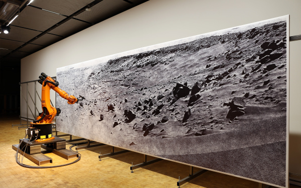An orange roboter draws a picture of a moonscape
