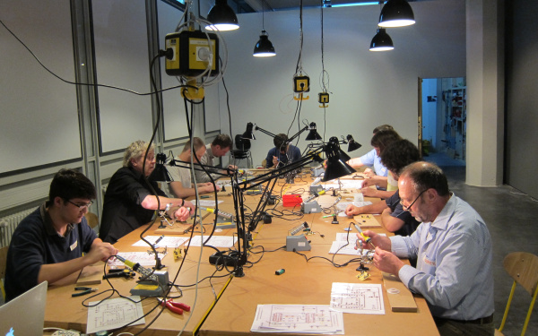 An einem riesigen Tisch sitzen viele Workshopteilnehmer, die mit Lötkolben arbeiten und es liegt allerlei Material kreuz und quer herum.