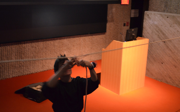 Das Foto zeigt eine Performerin im Vordergrund die an einem hängenden Seil ein weiteres befästigt. Sie trägt ein Nadelkissen um ihr linkes Handgelenk. Die schwarz gekleidete Person steht schräg vor dem Rednerpult auf dem orangenen Teppich.