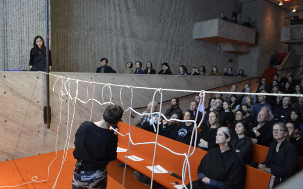 The photo shows a brutalist lecture hall with a view of the audience. The stage is cut. A large audience watches the performer form a net of ropes.