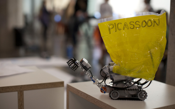 Auf einem Tisch steht ein kleiner Lego-Roboter, an dem ein gelbes Schild mit dem Wort "Picassoon" befestigt ist.