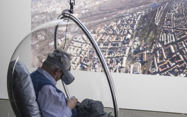 A visitor with VR glasses sits in a hanging semicircular armchair made of transparent plastic.