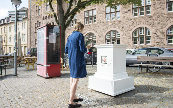 A woman in a blue dress stands in front of an empty white pedestal. The pedestal belongs to the AR artwork »The People's Monuments«.