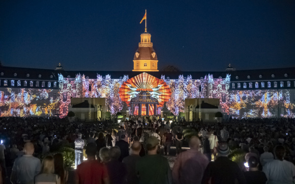 On the façade of Karlsruhe Castle, a huge digital shell emerges that slowly opens. A sea of colour surrounds it.