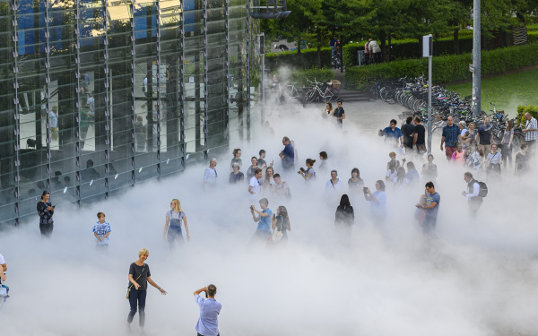 The photo shows a section of the glass cube of the ZKM in front of whose front right corner several visitors stand in the fog sculpture and take photos.