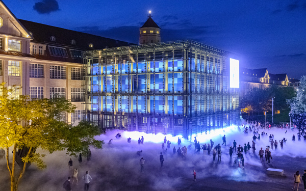 Das Foto ist bei Nacht mit einer Drohne aufgenommen. Es zeigt die Nebelskulptur und viele Silhouetten von BesucherInnen der KAMUNA 2019. Der Kubus und die Fassade des Gebäudes sind hell erleuchtet.  