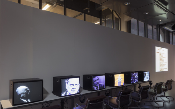 An einer Wand, ein langer Tisch mit sechs Fernsehern, auf denen Unterschiedliche Videos laufen. Dazu jeweils Kopfhörer und davor schwarze Stühle.