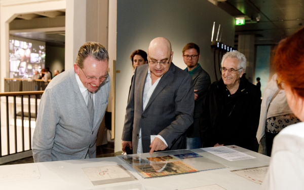 Michael Bielicky points explainingly to a newspaper article lying on a table. There are several people standing around him looking at him or the article.