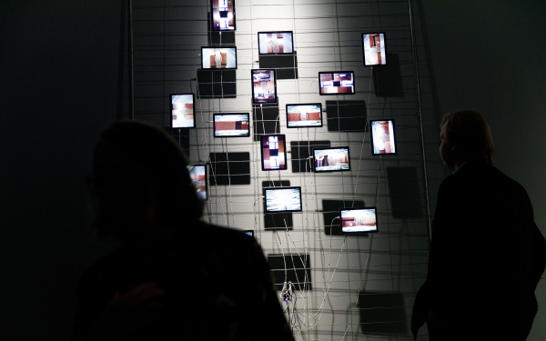 On a metal fence in front of a grey wall hang some tablets which are used as displays. The arrangement is striking, reminiscent of the classic Petersburg hanging. The wiring of the tablets is also notable. A person stands in front of the installation.