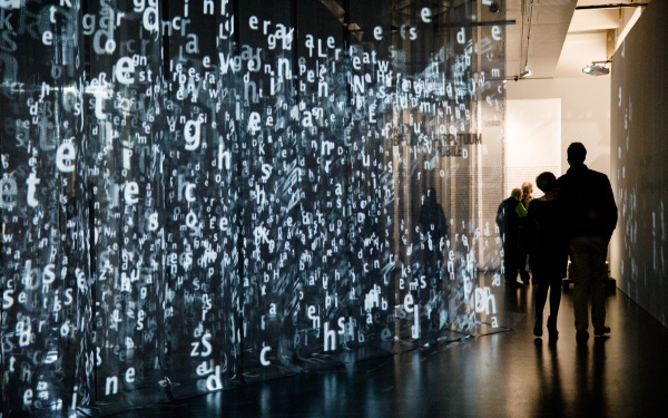 The picture shows a large number of hanging, transparent black curtains on which many luminous letters are projected. Two people walk past it, on the right.