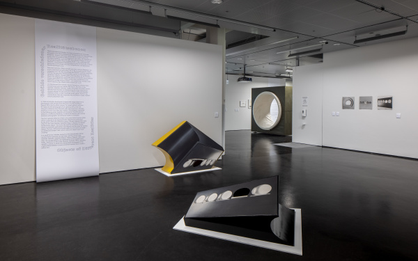 In the exhibition space, two sculptural works can be seen in the foreground. In the background, text is written on the wall and other works can be seen. 