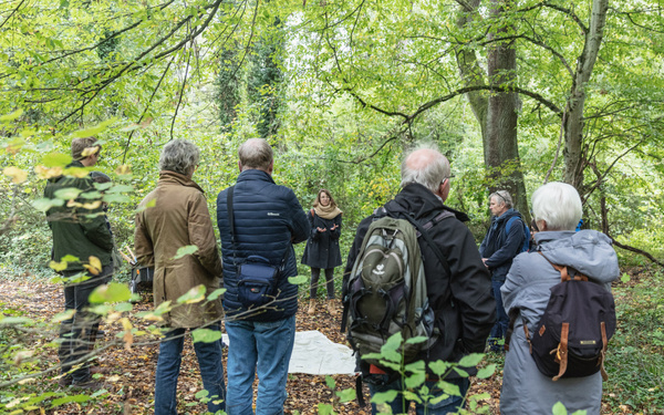 Eine Gruppe Menschen im Wald steht in Kreis um eine Frau, die spricht.