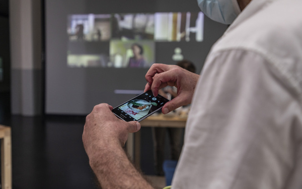 Eine Person fotografiert einen Teller mit Essen auf ihrem Smartphone. Im Hintergrund wird eine Videokonferenz auf die Wand projiziert
