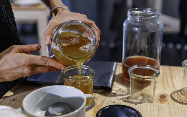 Aus einem Einmachglas wird eine Flüssigkeit in ein Glas auf einem Tische gefüllt. Auf dem Tisch stehen weitere Gläser und Schüsseln.