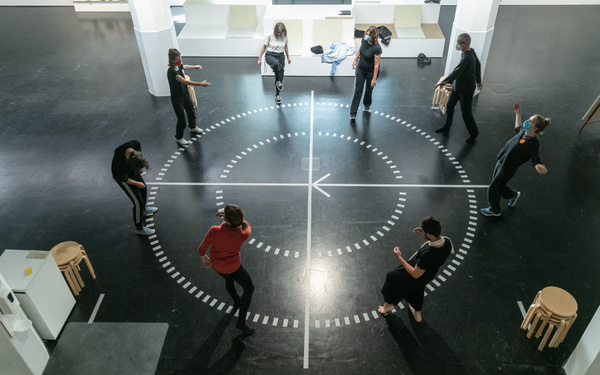 Eine Gruppe Menschen steht im Kreis auf einem runden Diagramm auf einem Hallenboden. Die Menschen scheinen in Bewegung zu sein.