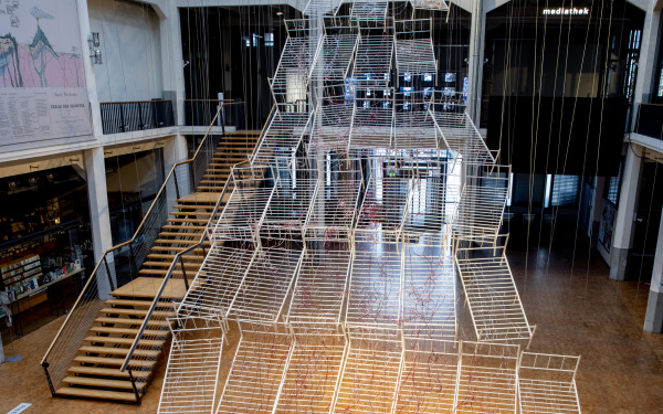 Installationsansicht von Chiharu Shiota`s Werk »Connected to Life«. Zu sehen sind aneinandergereihte, miteinander verbundene Bettgestelle, durchzogen von roten Schläuchen, die sich immer weiter vom Boden abheben und gemeinsam eine Art Weg aufwärts bilden.