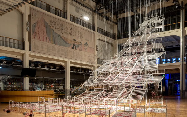 Installationsansicht von Chiaru Shiotas »Connected to Life«. Zu sehen sind Krankenhausbettengestelle, die pyramidenförmig von der Decke hängen. Durchzogen sind diese von Schläuchen, durch die rote Farbe fließt.