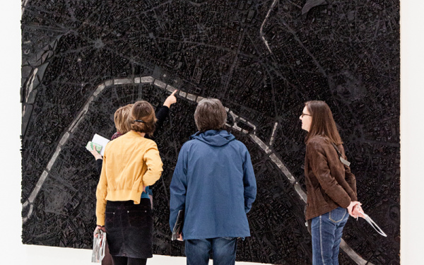 Zu sehen sind vier Personen, die vor einem dreidimensionalen, an der Wand hängenden Bild befinden. Das Bild ähnelt einer Stattkarte, die gänzlich schwarz eingefärbt und von wenigen weißen Linien durchzogen ist