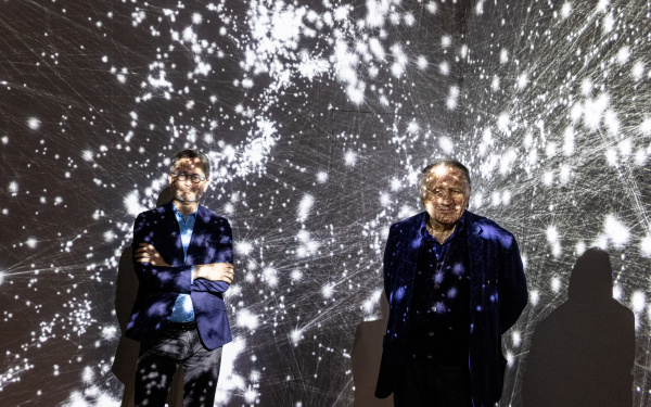 Two men stand in the exhibition room in front of a projection that shows the cosmos as a network. They are covered by the projection. The left man is the scientist Albert-Lázsló Barabási. The right man is Peter Weibel. Both are smiling.
