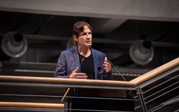 The artist Jean-Remy stands on the ZKM foyer stairs at the railing behind a lectern and speaks. His gestures accompany his speaking.