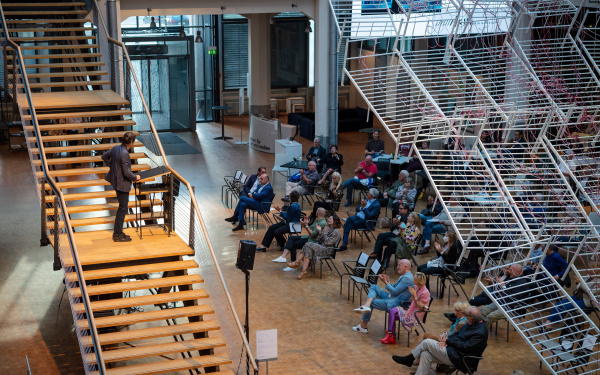 The artist Jean-Remy speaks to the visitors on the stairs in the ZKM foyer during the opening of the exhibition. The visitors sit on chairs at a distance and listen. Above them hangs an installation of bed frames
