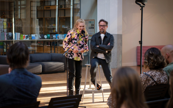 Die SchauspielerInnen Anna Loos und Jan Josef Liefers sind hinter einem Mikrofon. Anna Loos spricht, Jan Josef Liefers sitzt auf einem Hocker, die Arme verschränkt und hört zu.